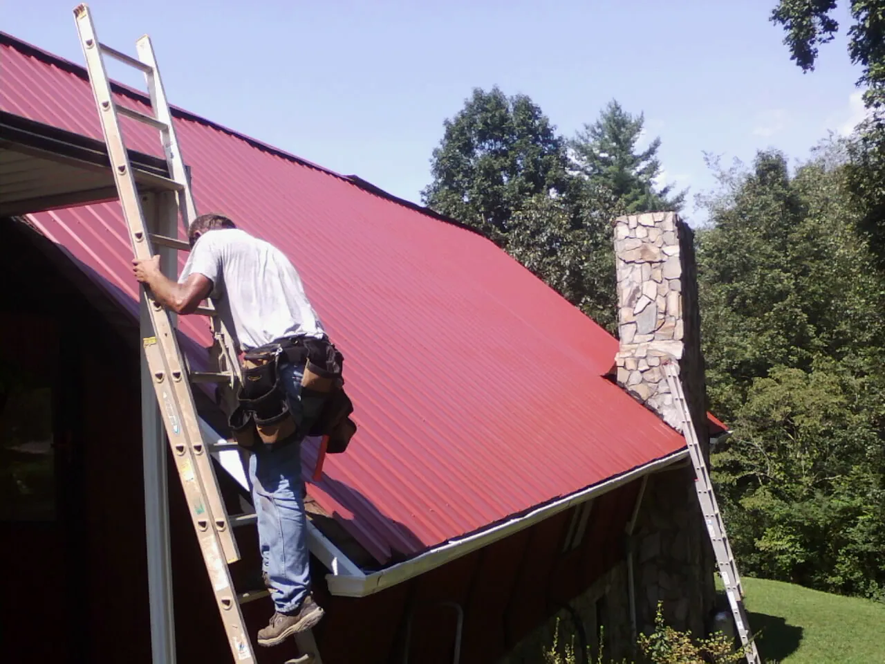 Roofer on a ladder beside a red metal roof and stone chimney — Soffit & Fascia Repair by Roofing Brothers in Cheyenne